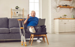 An eldery man, with his right leg in a cast, meets with a telerehabilitation doctor via video conference.