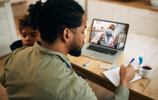 Father sits with son at a desk as the father talks to doctor via laptop.