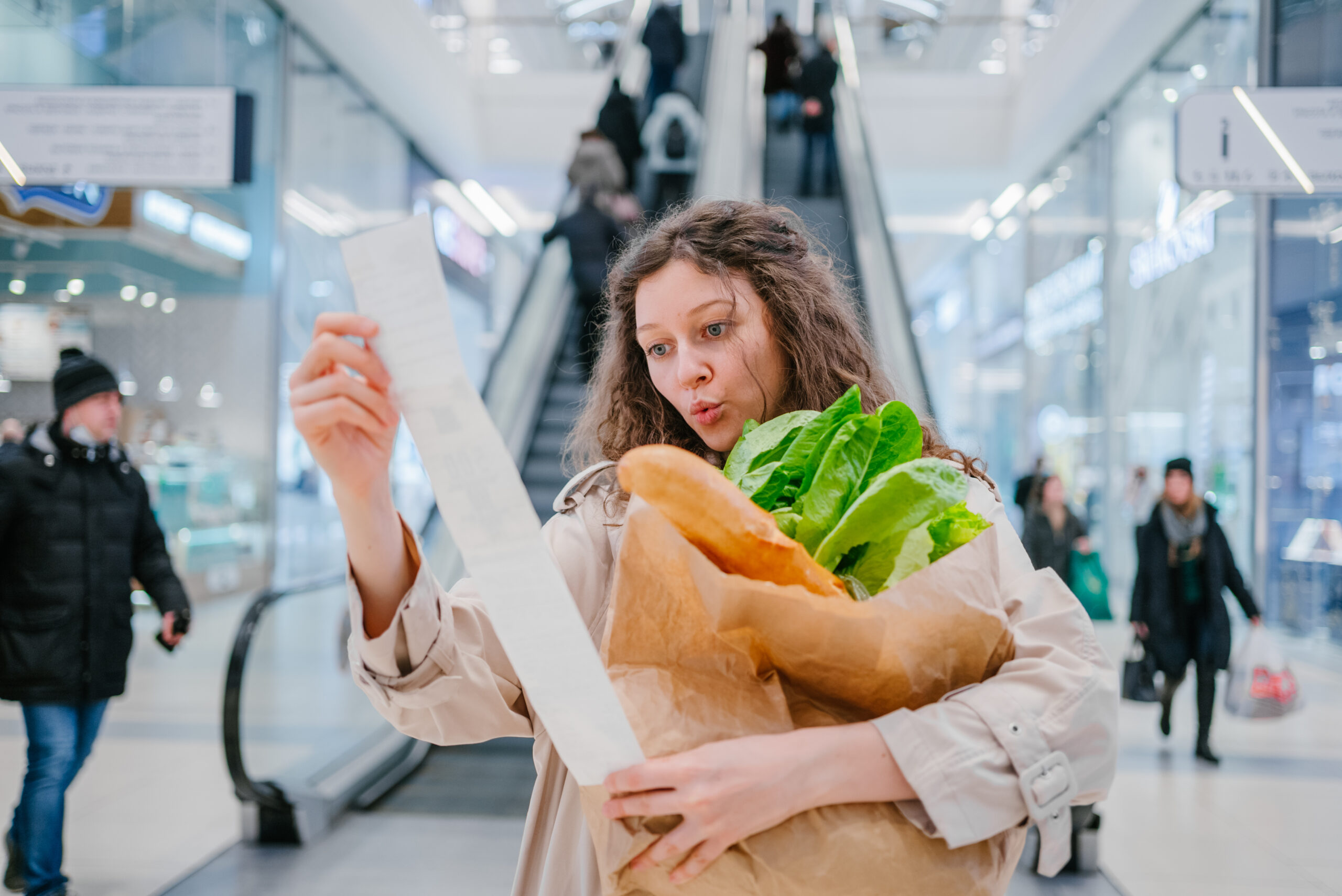 Rising food prices in part due to inflation. Surprised woman looking into a paper check at the mall, holding a paper bag with fresh herbs and a baguette.