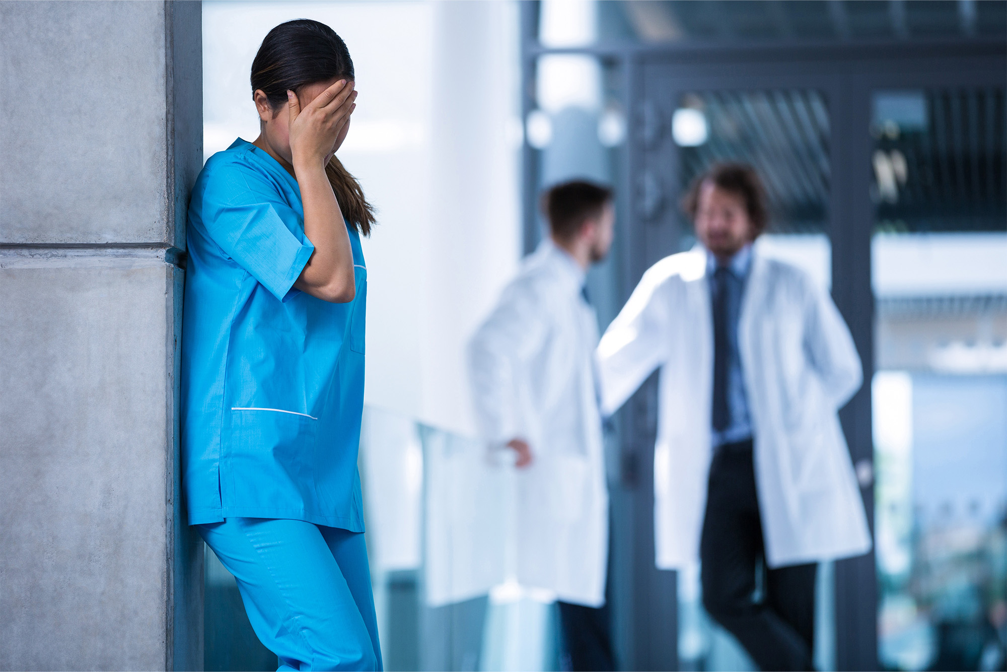 Nurse experiencing stress while two doctors converse in the background.