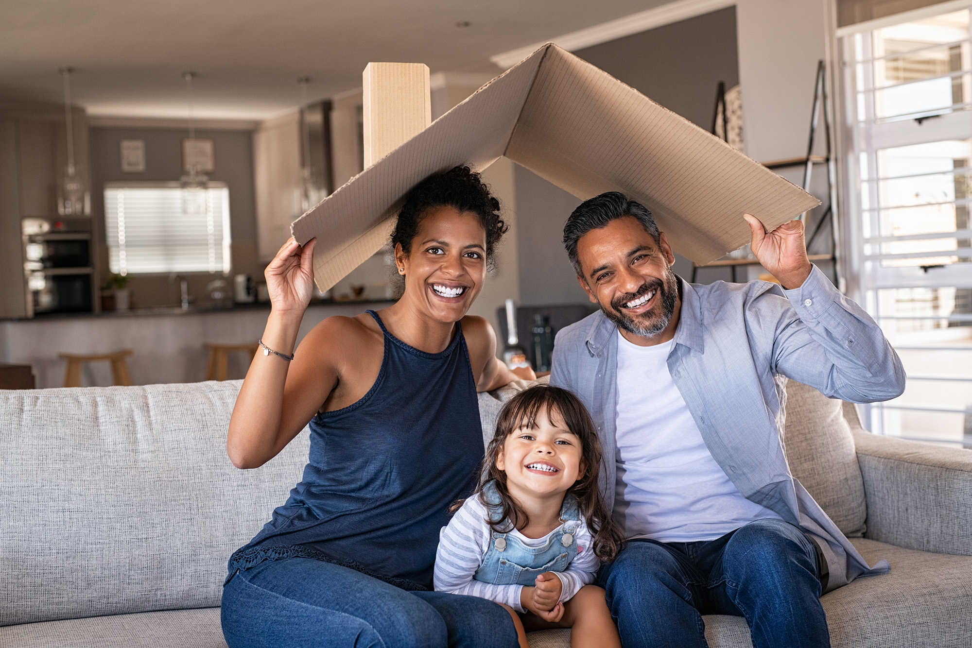 A family of three sitting on a couch with a cardboard box symbolizing a roof over their heads.