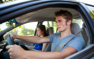 A teen driver in the driver's seat while a female passenger sits on her cell phone.