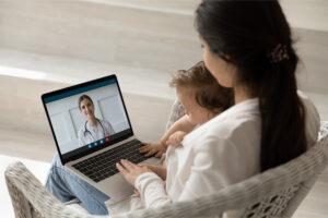 A woman sitting in a chair, holding a young child, while communicating with a doctor via conference call.