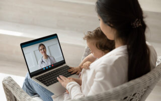 A woman sitting in a chair, holding a young child, while communicating with a doctor via conference call.