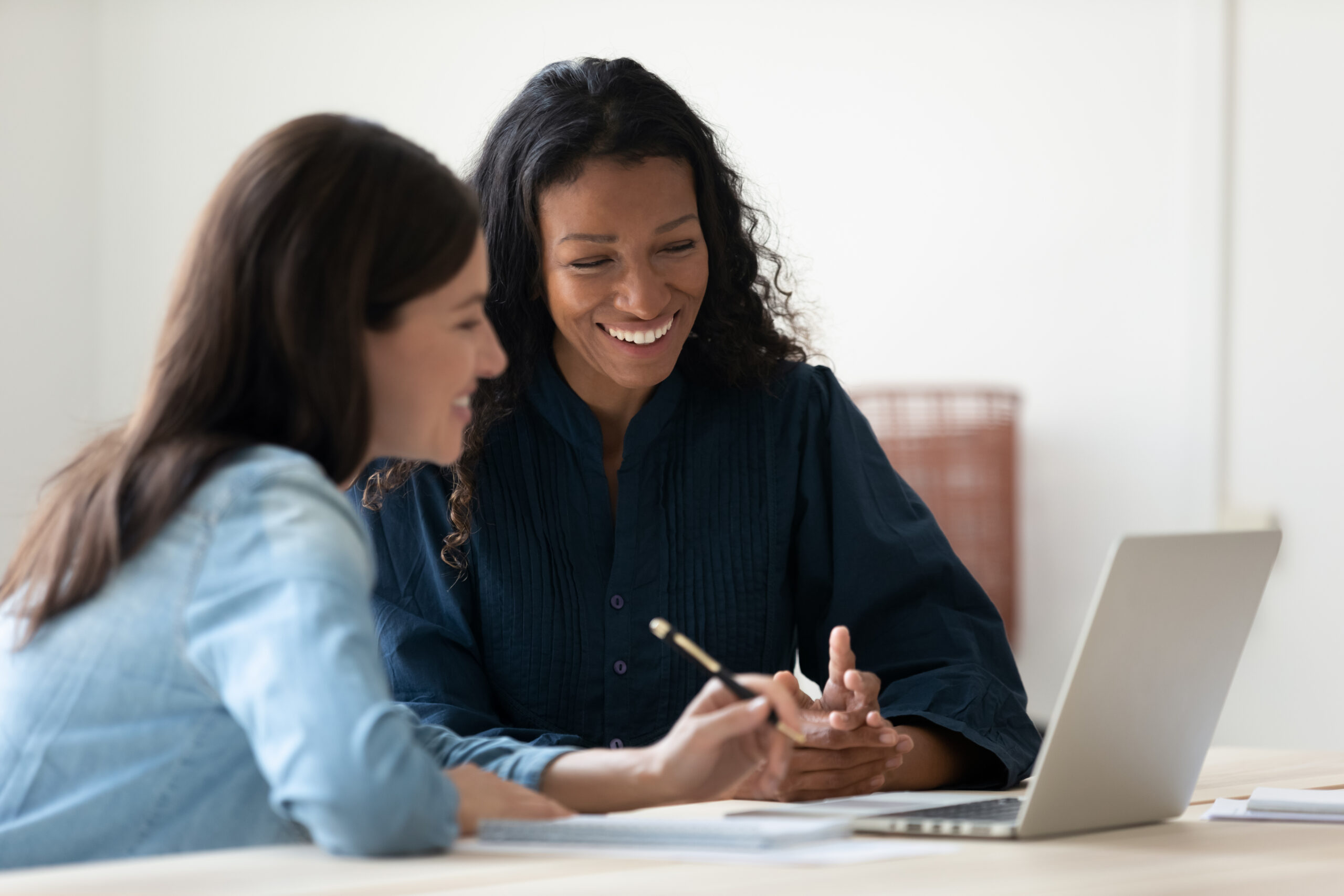 Close up happy young african american businesswoman using laptop with laughing female mentor in coworking boardroom at meeting. Smiling diverse woman managers talking about employee benefits options