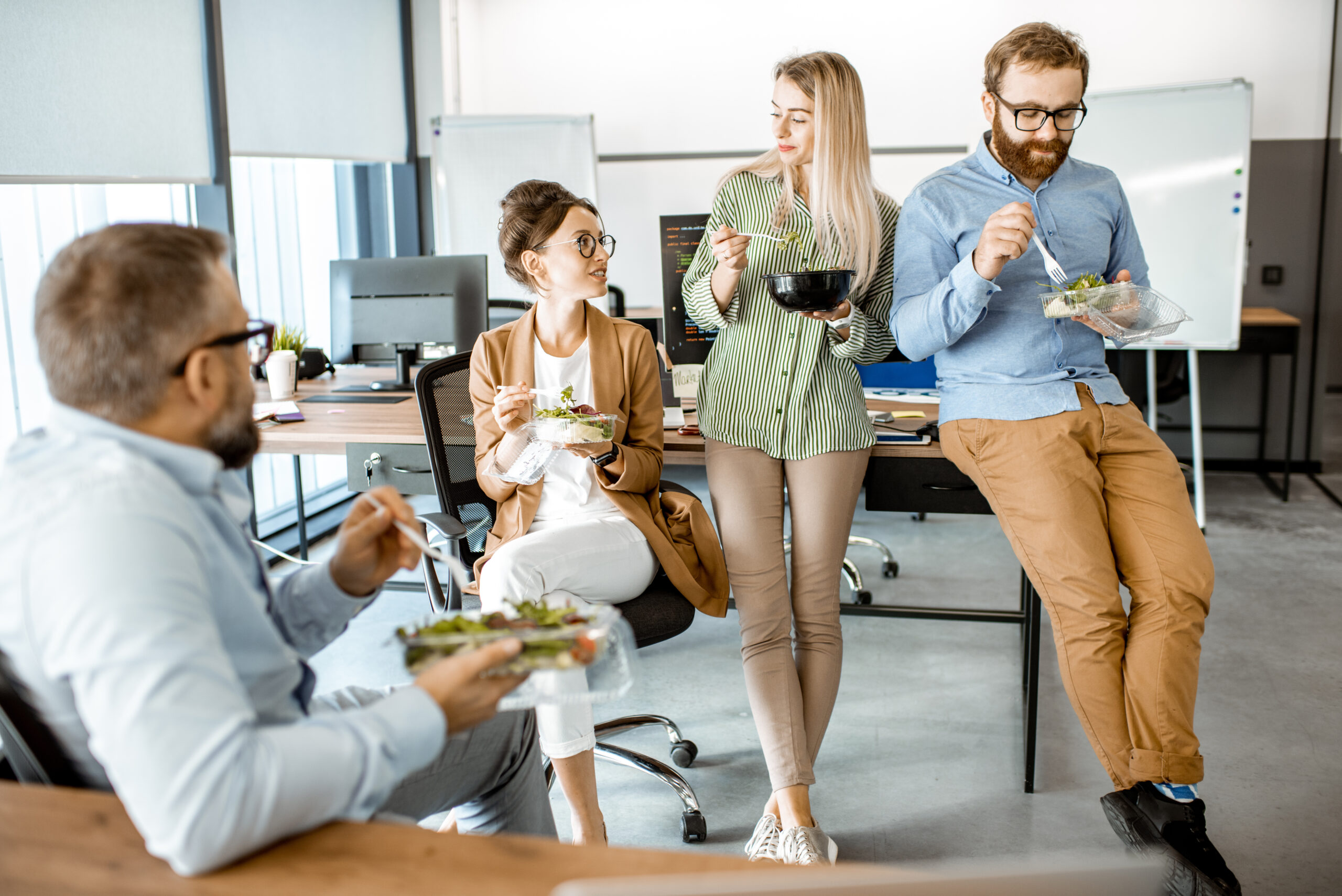 Attraction and Retention Trends: Group of diverse colleagues eating takeaway salad, sitting together and having fun during a lunchtime in the office.