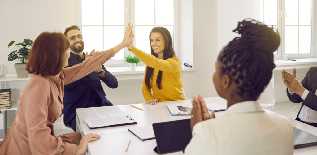 A group of colleagues sitting around a table as two women high-five each other, signifying a healthy work environment.
