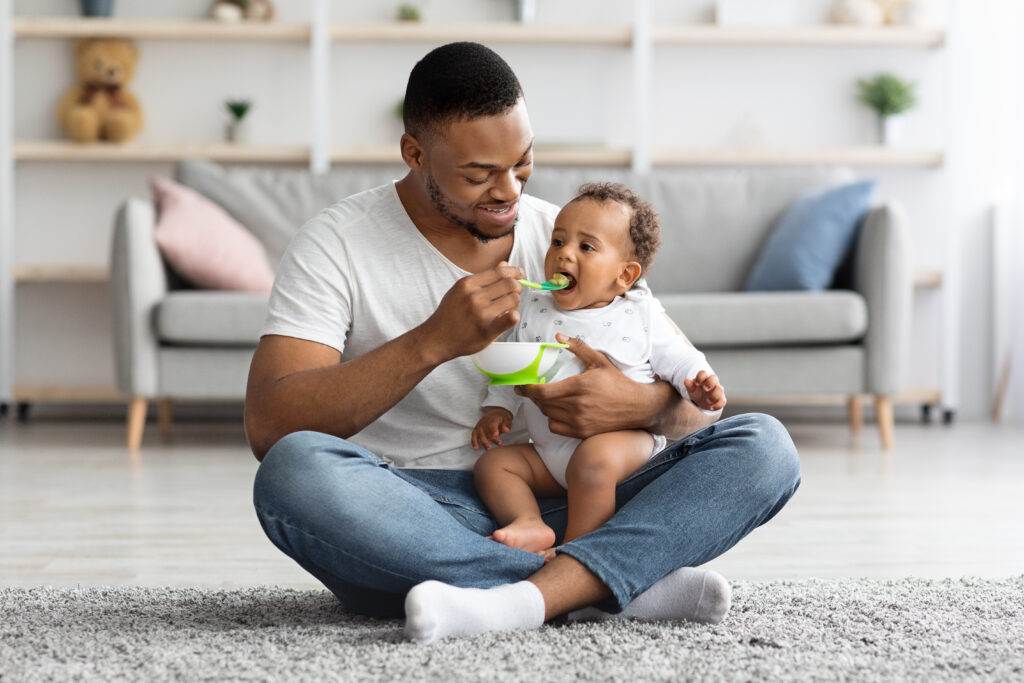 Dad Feeding His Infant Baby From Spoon While Sitting Together On Carpet In Living Room