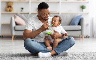 Dad Feeding His Infant Baby From Spoon While Sitting Together On Carpet In Living Room