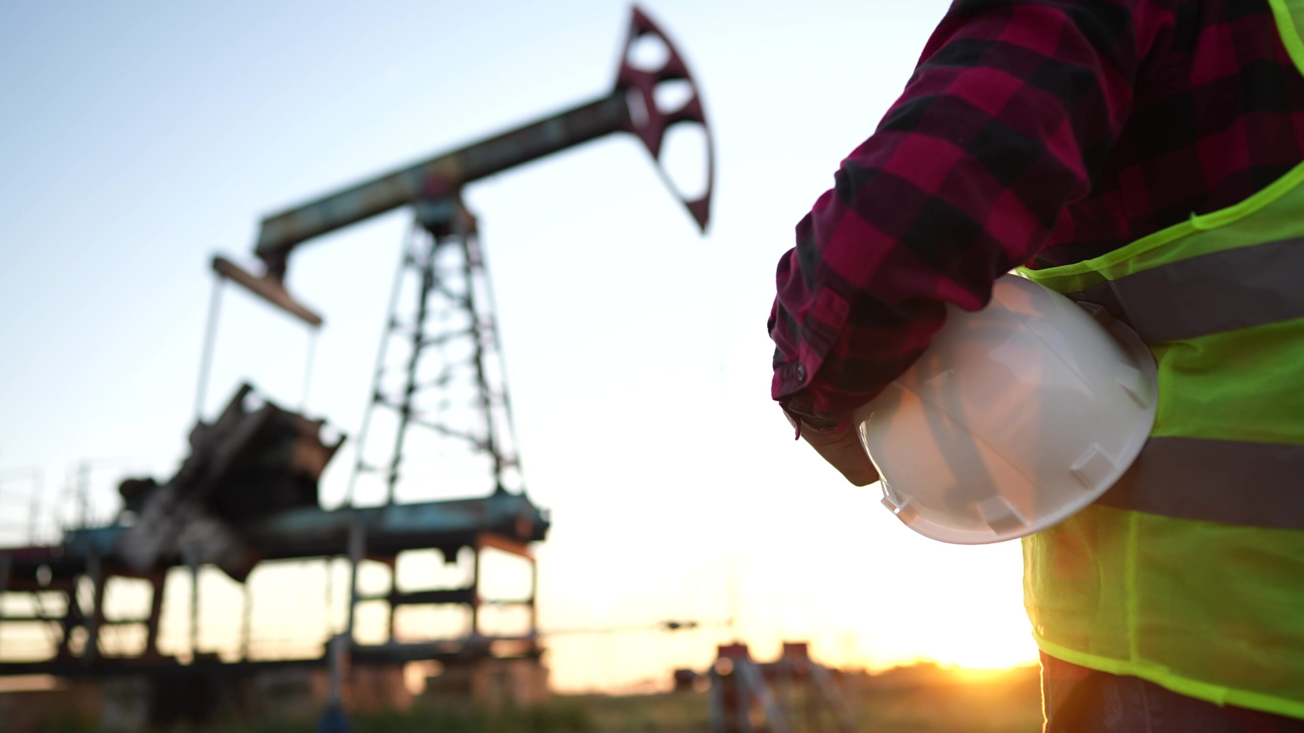 oil production. a worker holding protective hard hat at sunset in the background an oil pump.
