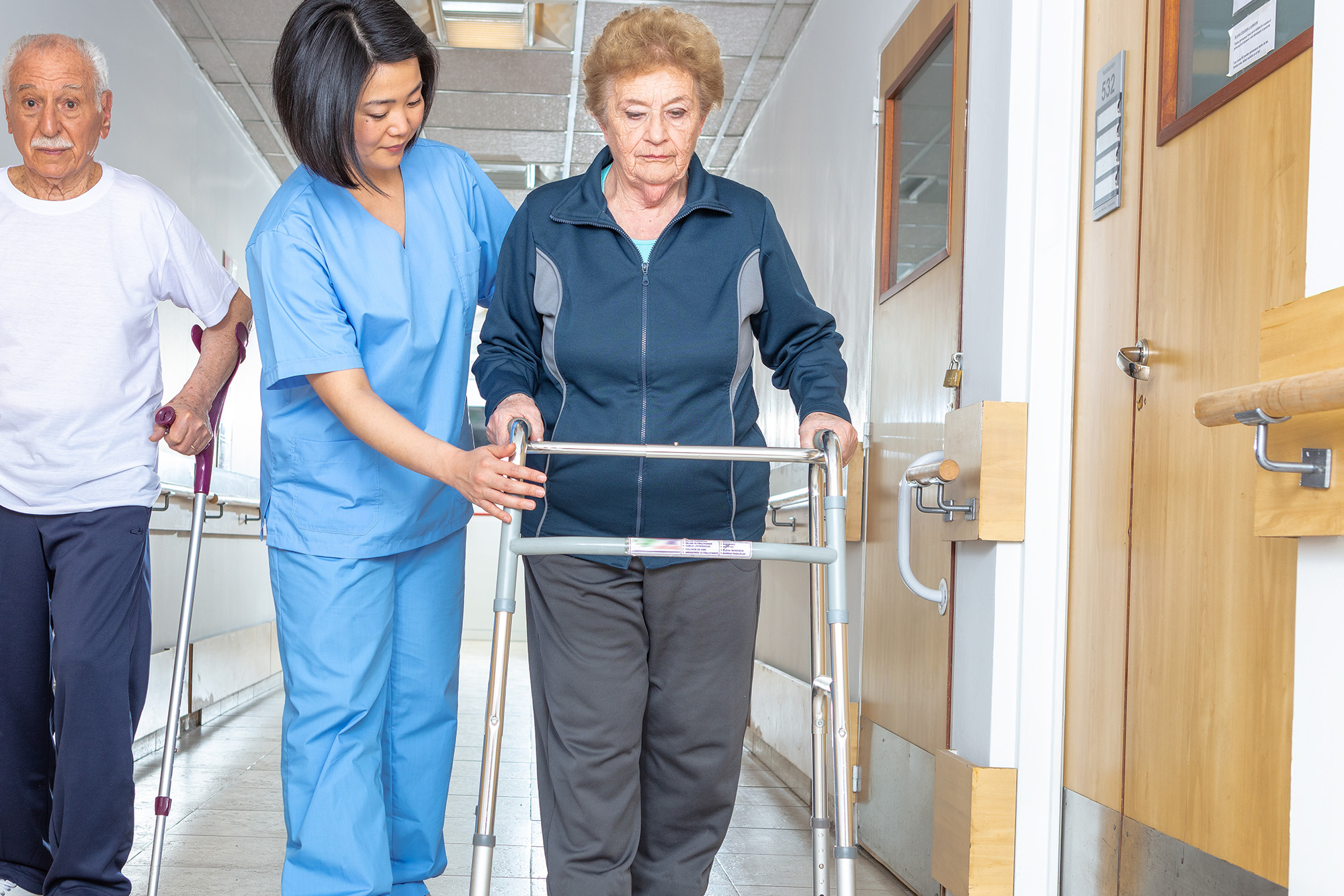 patient transfers: doctor helping elder woman with walker and man in hospital aisle.