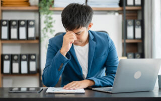 Rising Mental Health Problems at Work: a young business professional sitting at his desk with his hand over his face as he appears stressed.