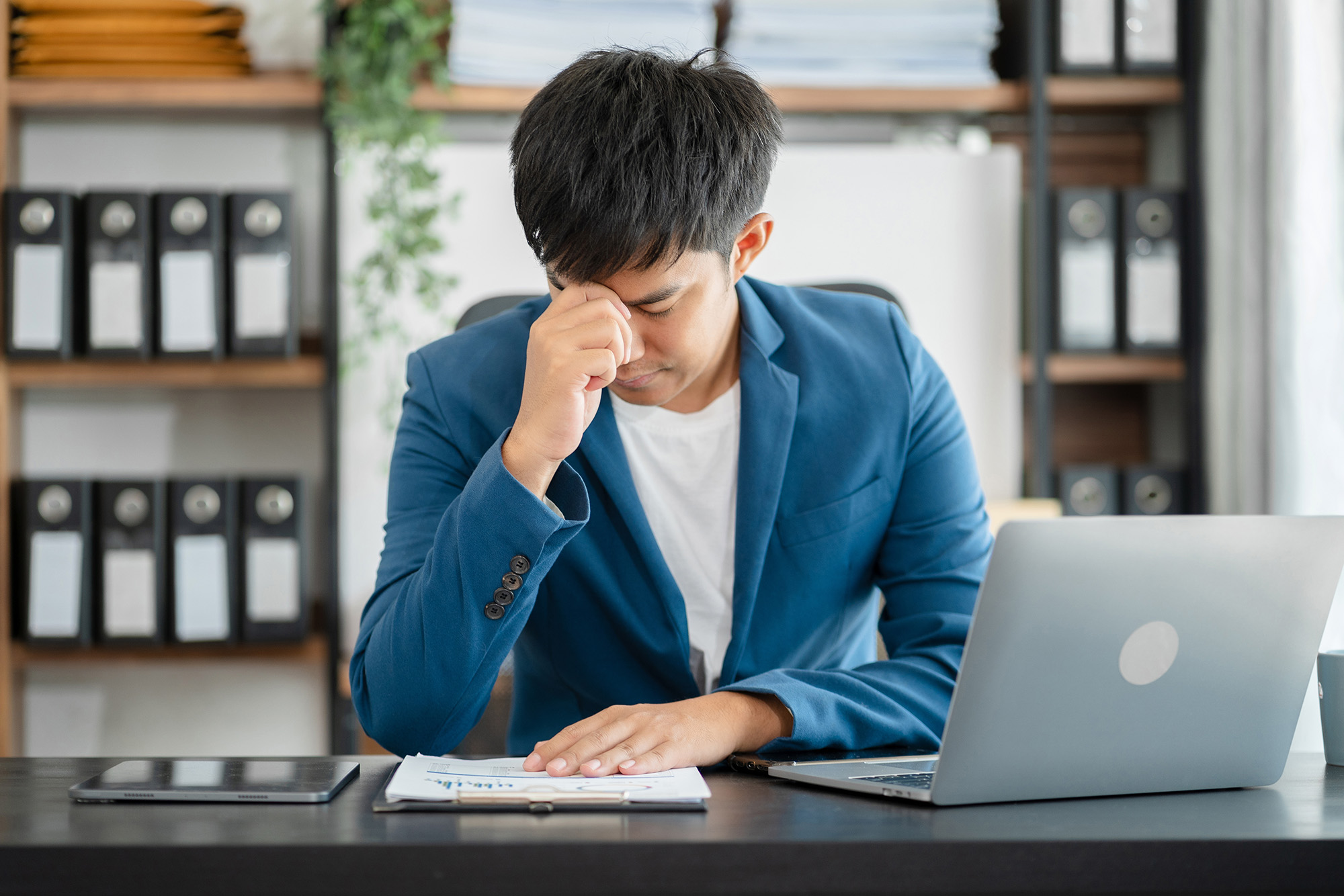 Rising Mental Health Problems at Work: a young business professional sitting at his desk with his hand over his face as he appears stressed.