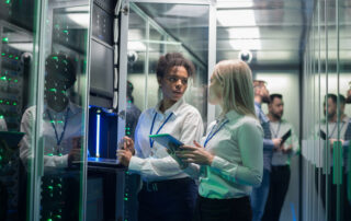 Cybersecurity Awareness Month: Medium shot of two women working in a data center with rows of server racks and checking the equipment and discussing their work