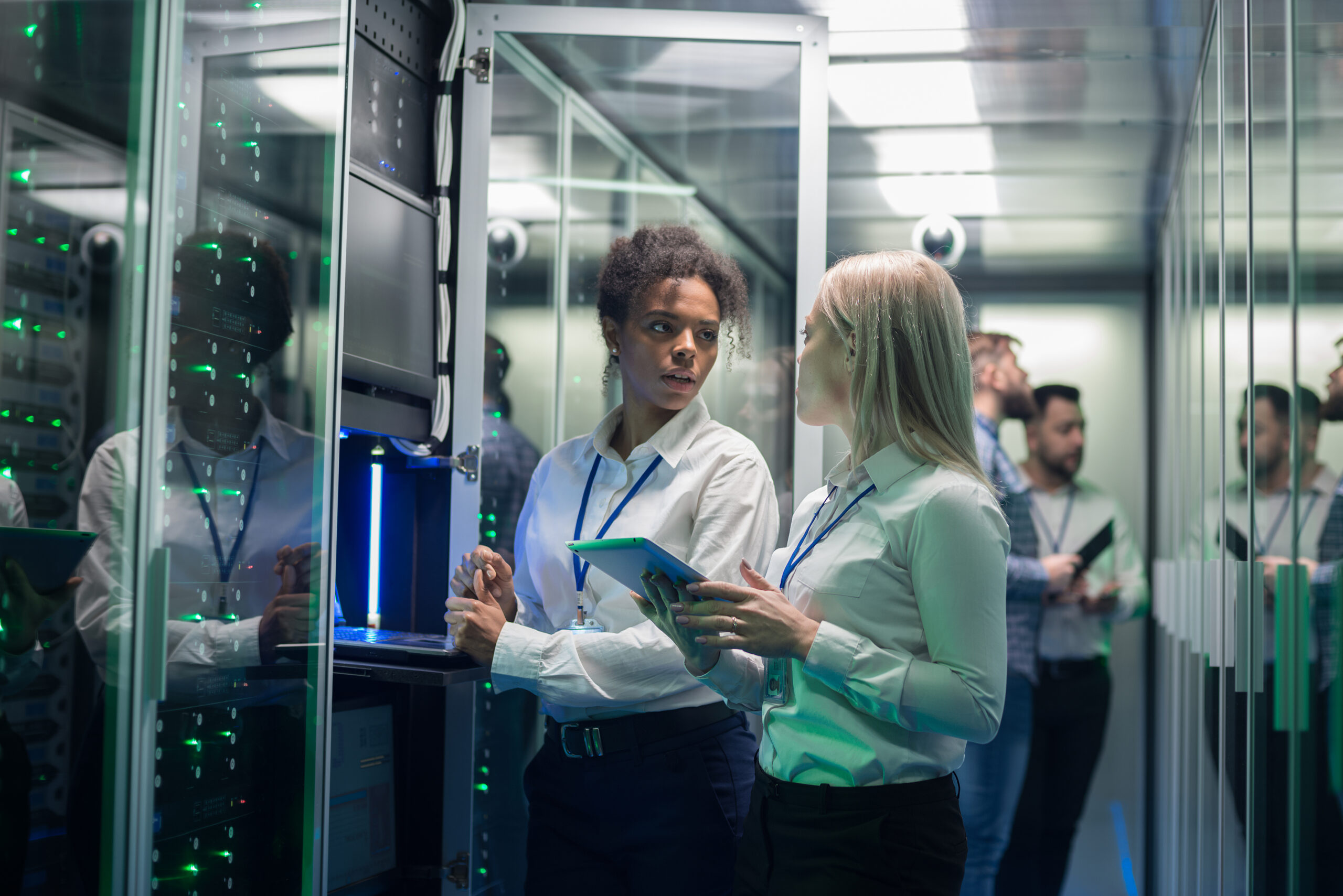 Cybersecurity Awareness Month: Medium shot of two women working in a data center with rows of server racks and checking the equipment and discussing their work