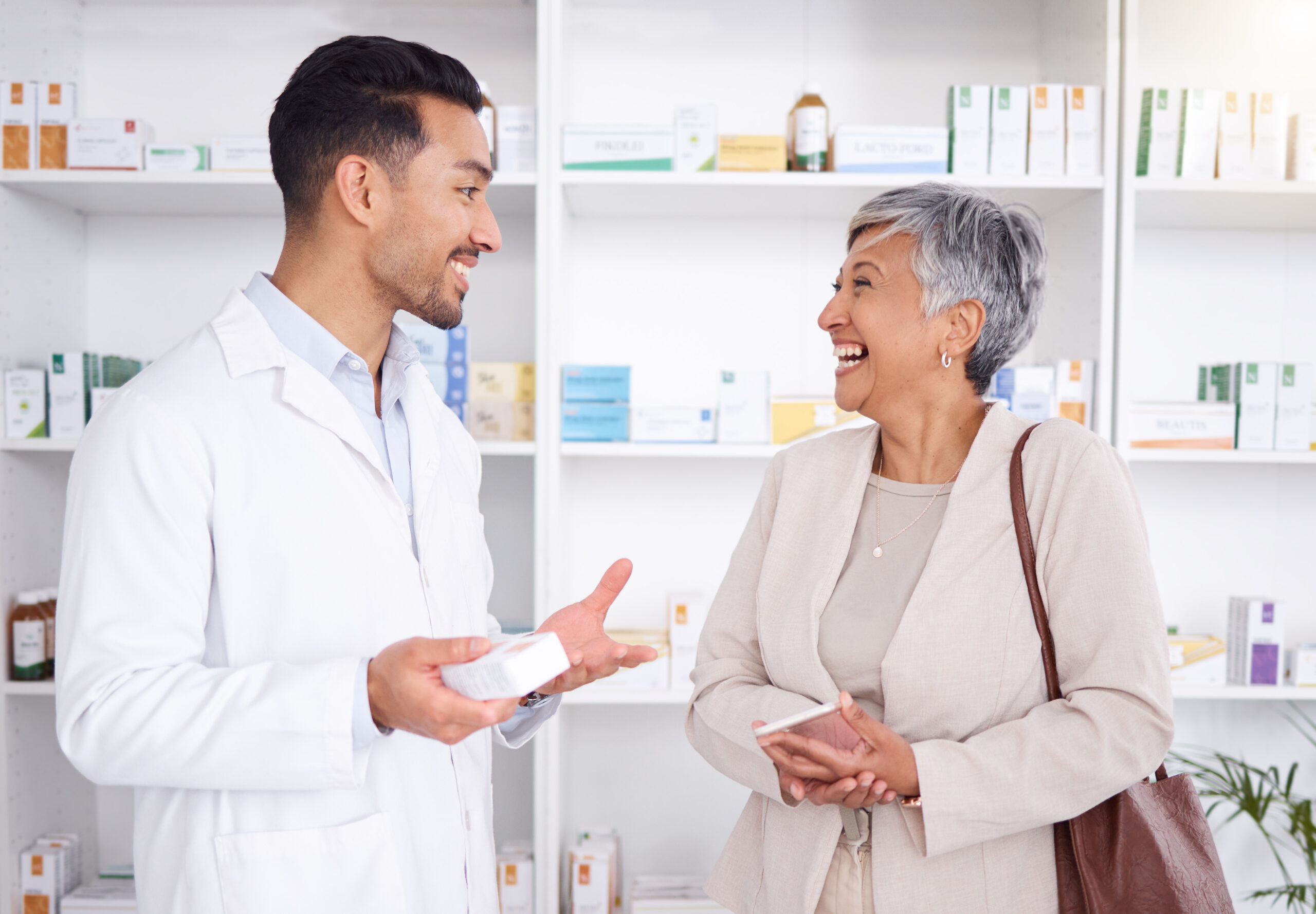 4 Key Trends Driving Employer Healthcare Costs in 2024: Pharmacist man, senior woman and talking with box, phone or funny in store for prescription, health or help. Young pharmacy manager, elderly patient and comic chat for care with customer experience.