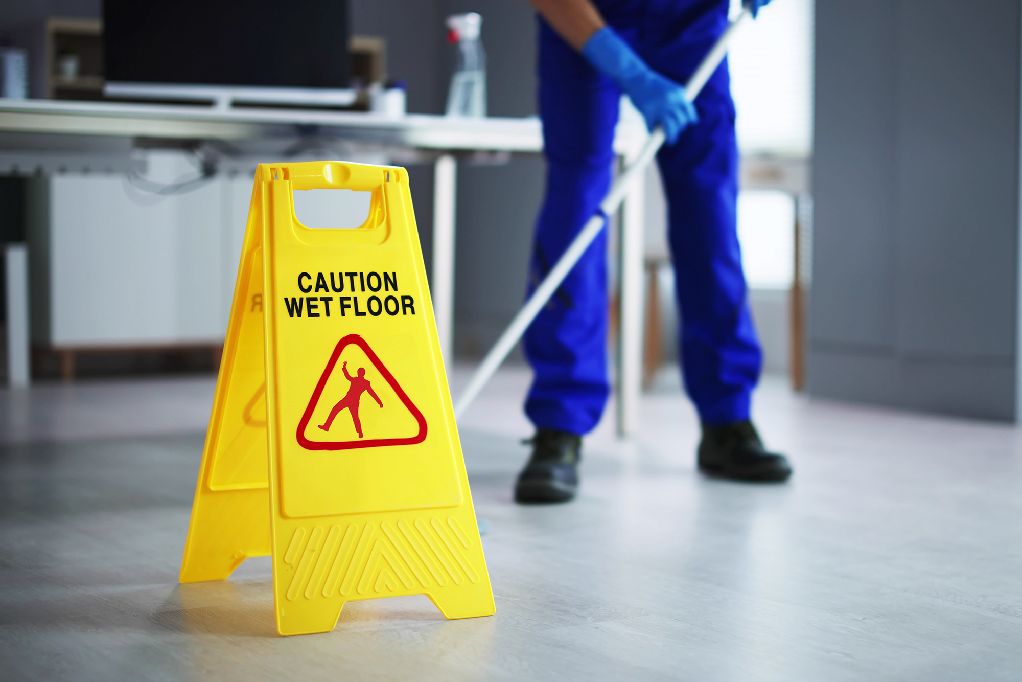 5 Tips for Preventing Slips, Trips, and Falls on School Premises: A janitor cleaning up a classroom with a mop. There is a Caution Wet Floor sign in the foreground.