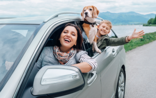 Tips for Auto Insurance Buyers in 2024: A mother in the driver's seat of a sedan with her young child and pet dog looking out the passenger window.