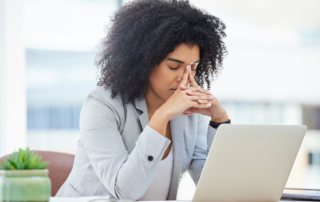 Supporting Employees During Mental Health Awareness Month and Beyond: A stressed out female colleague sitting at her desk with her head in her hands.