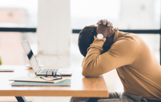 Preventing Burnout in Working Parents Helps Employers: A male with his head in his hands in front of his computer.