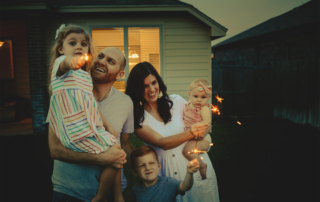 Firework Safety: A mother and father holding two children while the third is playing with sparklers.