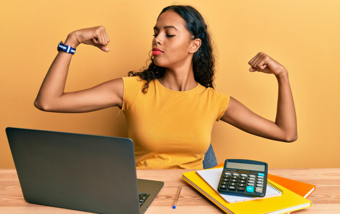 rethinking employee benefits: a professionally-dressed business woman flexing her biceps while sitting at her laptop.